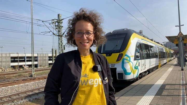 Frau steht am Bahngleis. Im Hintergrund steht ein bwegt-Zug mit angepasstem Design - der Batteriezug.