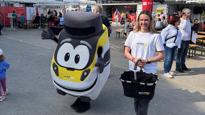 Die bwegt-Bine und eine Frau mit Korb begrüßen Besucher an einem Stand.
