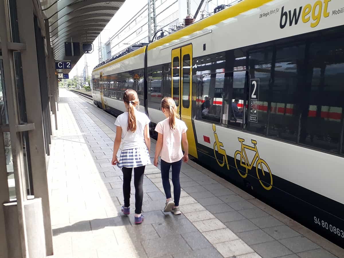 Zwei Mädchen stehen mit dem Rücken zur Kamera am Bahnsteig neben einem Zug.