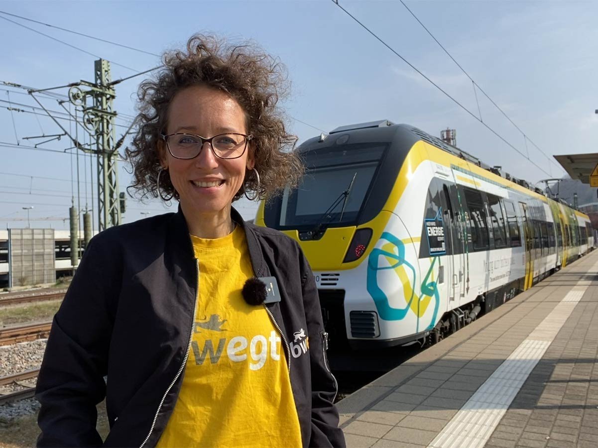 Frau mit lockigem Haar steht am Bahnsteig vor einem Batteriezug.