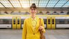 Eine Frau in gelber Kleidung steht an einem Bahnhof. Hinter ihr fährt ein bwegt-Zug vorbei.