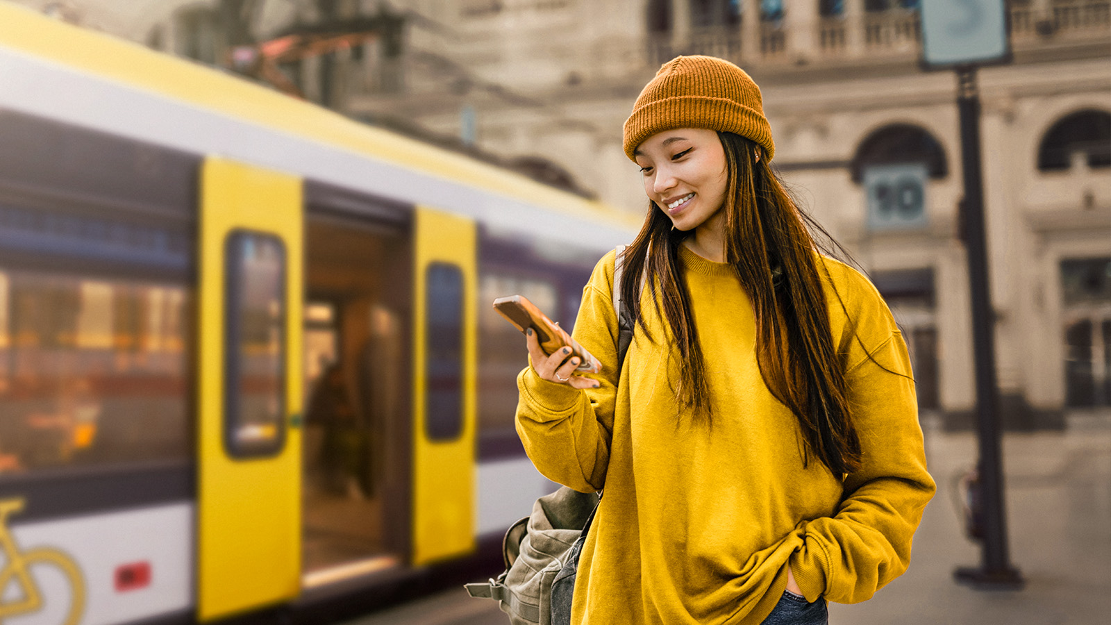 Eine junge Frau in gelber Kleidung und Mütze steht vor einer gelben Straßenbahn und schaut auf ihr Handy.