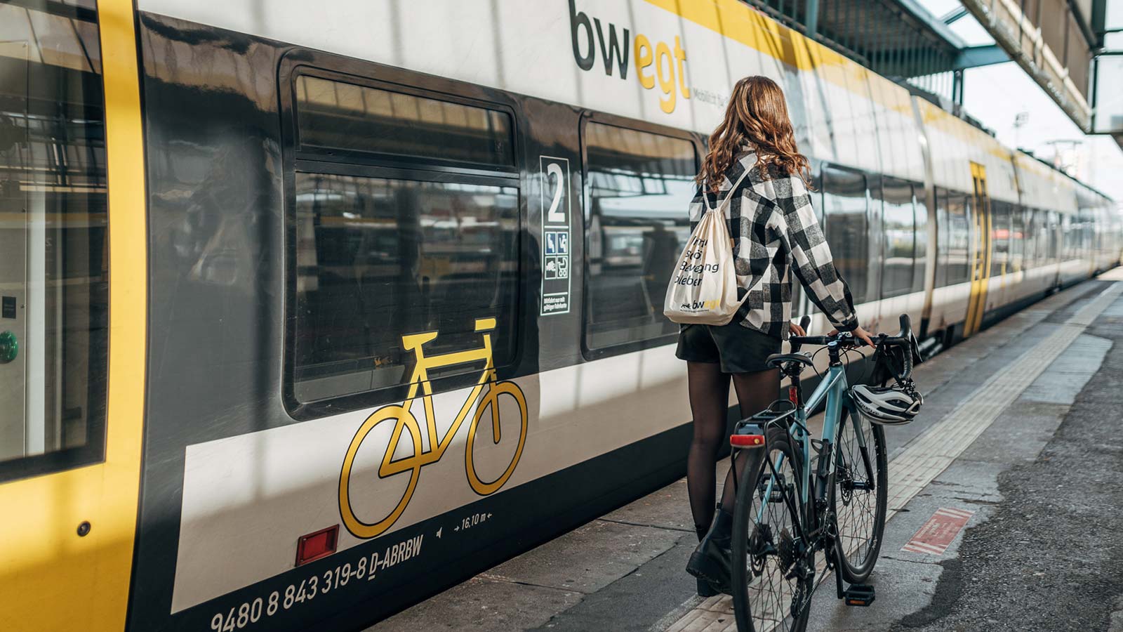 Mädchen läuft mit ihrem Fahrrad auf einem Bahngleis neben einer Bahn her.