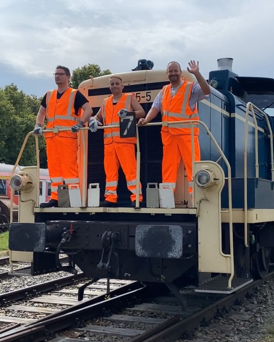 Drei Lokführer mit Warnweste stehen auf einer Bahn und winken.