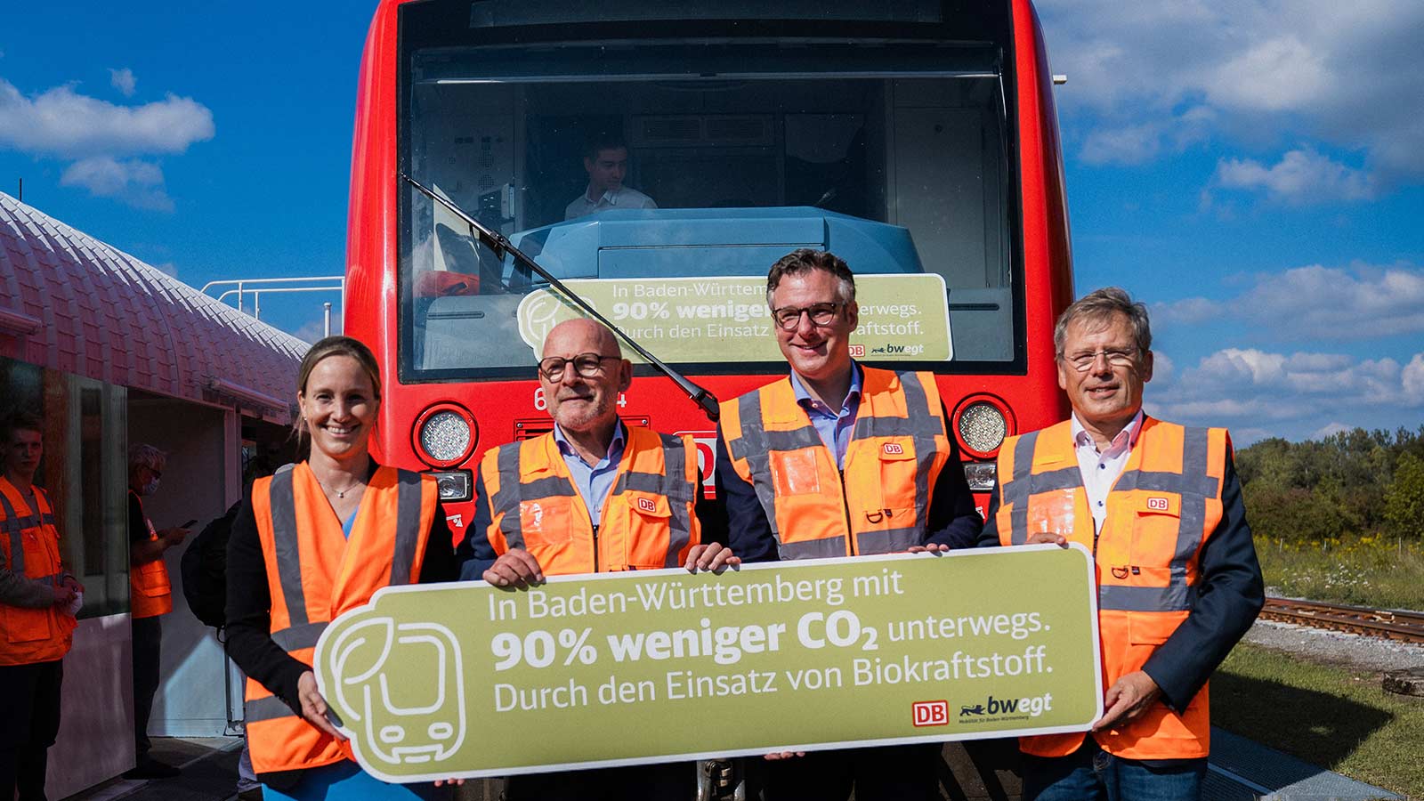 Drei Männer und eine Frau in Warnweste stehen mit einem Schild vor einem Zug. Auf dem Schild steht, dass durch Biokraftstoff 90 Prozent CO2 gespart wird.