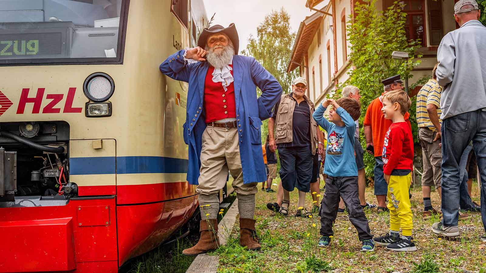 Ein älterer Mann im Räuber-Outfit lehnt an einer Bahn. Um ihn herum stehen mehrere Kinder.