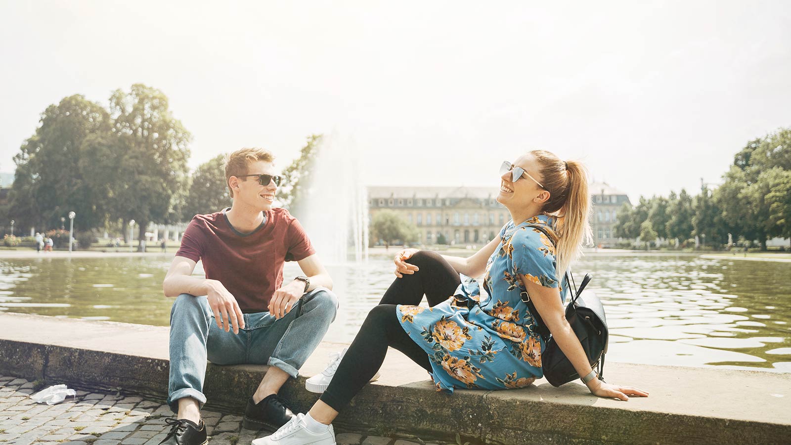 Junger Mann und Frau sitzen in der Stadt an einem Teich mit Springbrunnen.