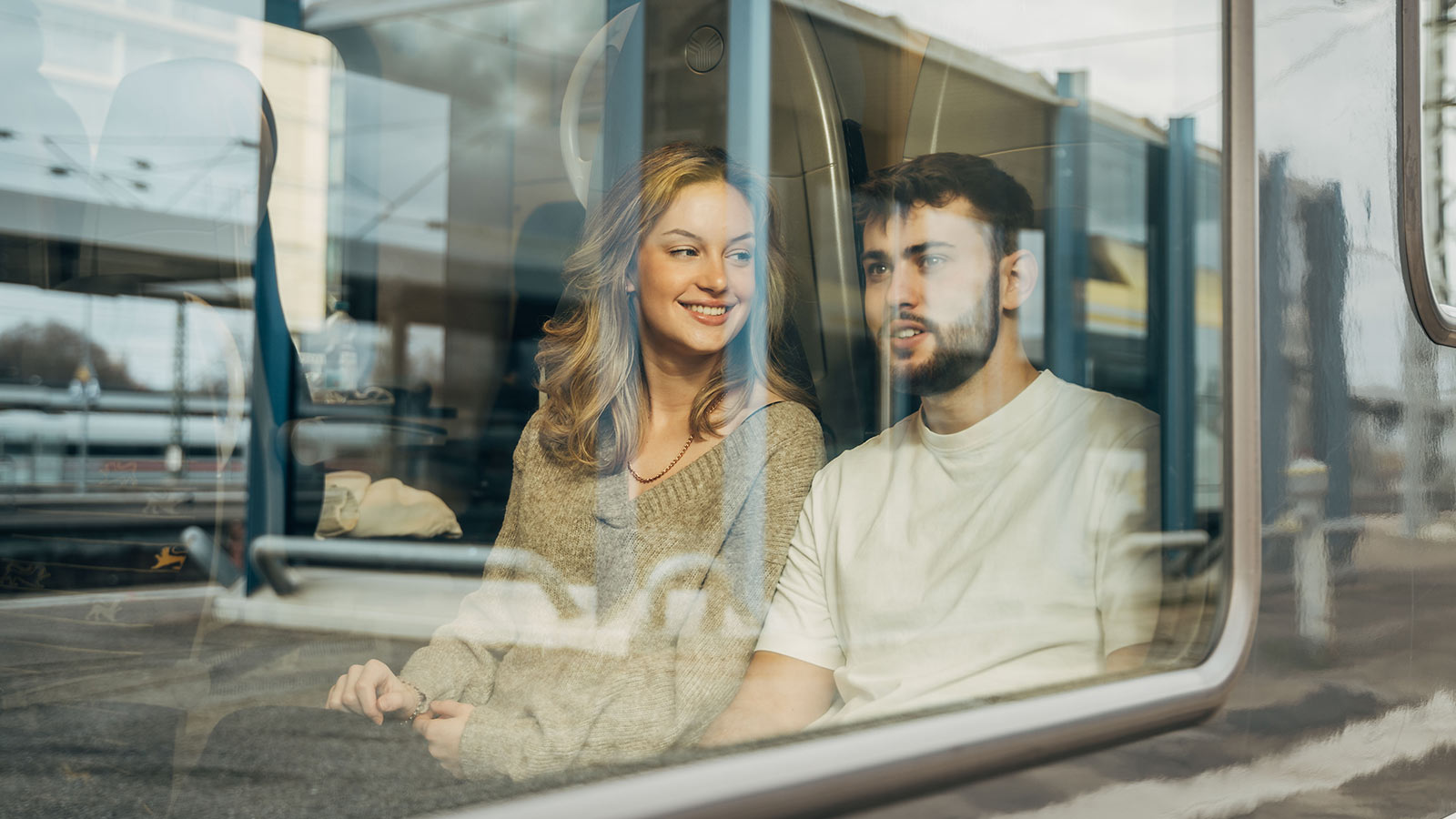 Junge Frau und Mann sitzen im Zug. Sie sind durch das Fenster zu sehen.